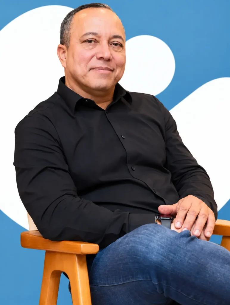 Retrato de Luciano Bravo sentado, usando camisa preta e calça jeans.